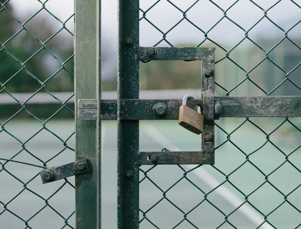 pexels-photo-16028000-16028000 Close-up of a locked gate with chain link fence at a tennis court outdoors.